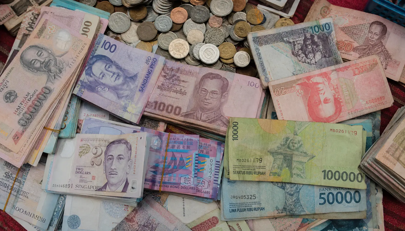 A market-table flat-lay of mixed Asian banknotes and coins &mdash; Vietnamese dong, Thai baht, Indonesian rupiah, Philippine peso, Singapore dollars and more
