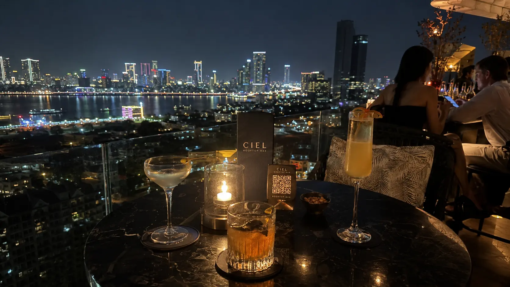 Phnom Penh rooftop bar at night with patrons leaning against the railing while the city's high-rise lights and the Mekong river spread out below