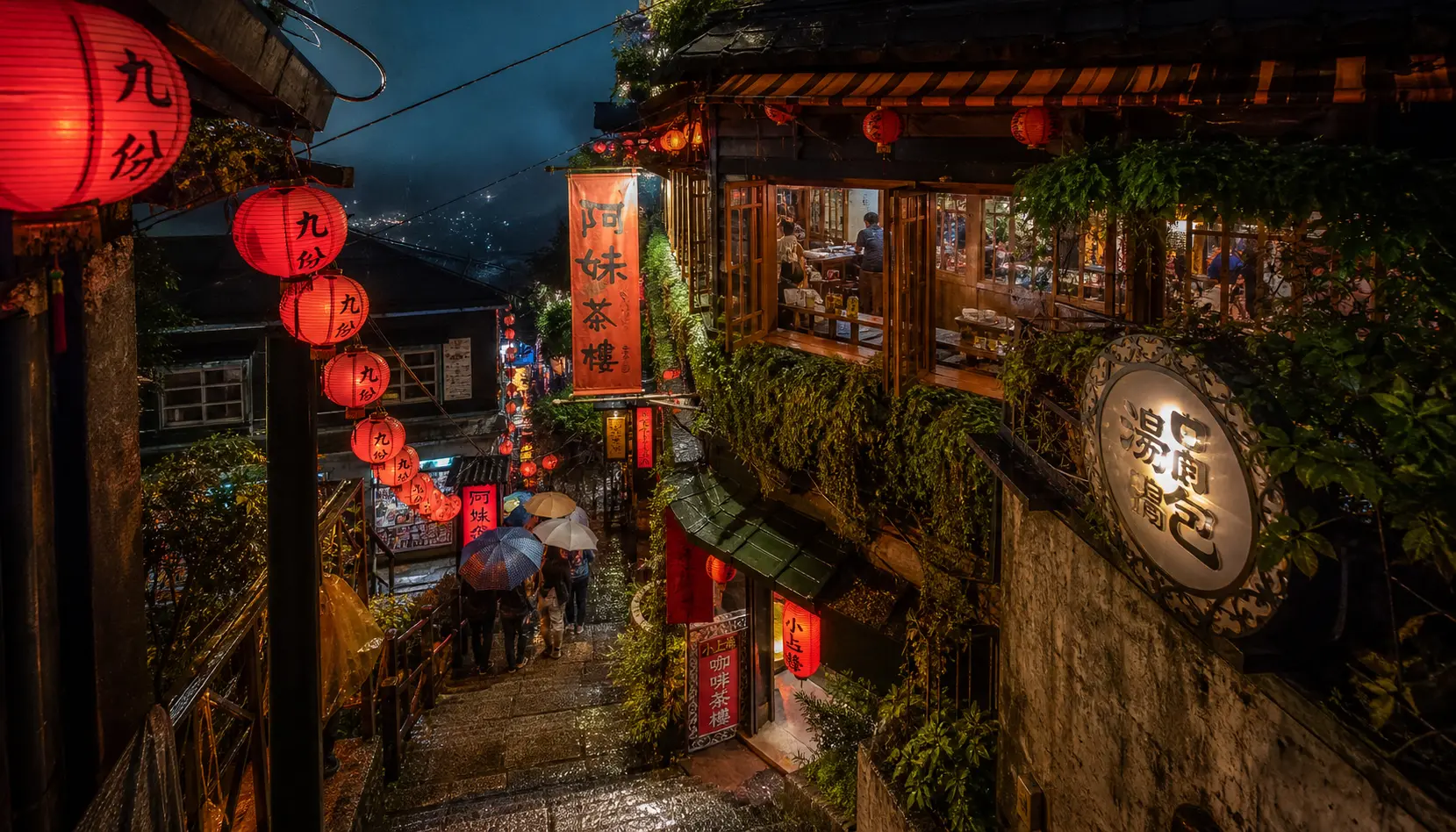Jiufen Old Street at night in light rain &mdash; red lanterns glowing over the stepped lane and the A-Mei teahouse, umbrellas in the crowd